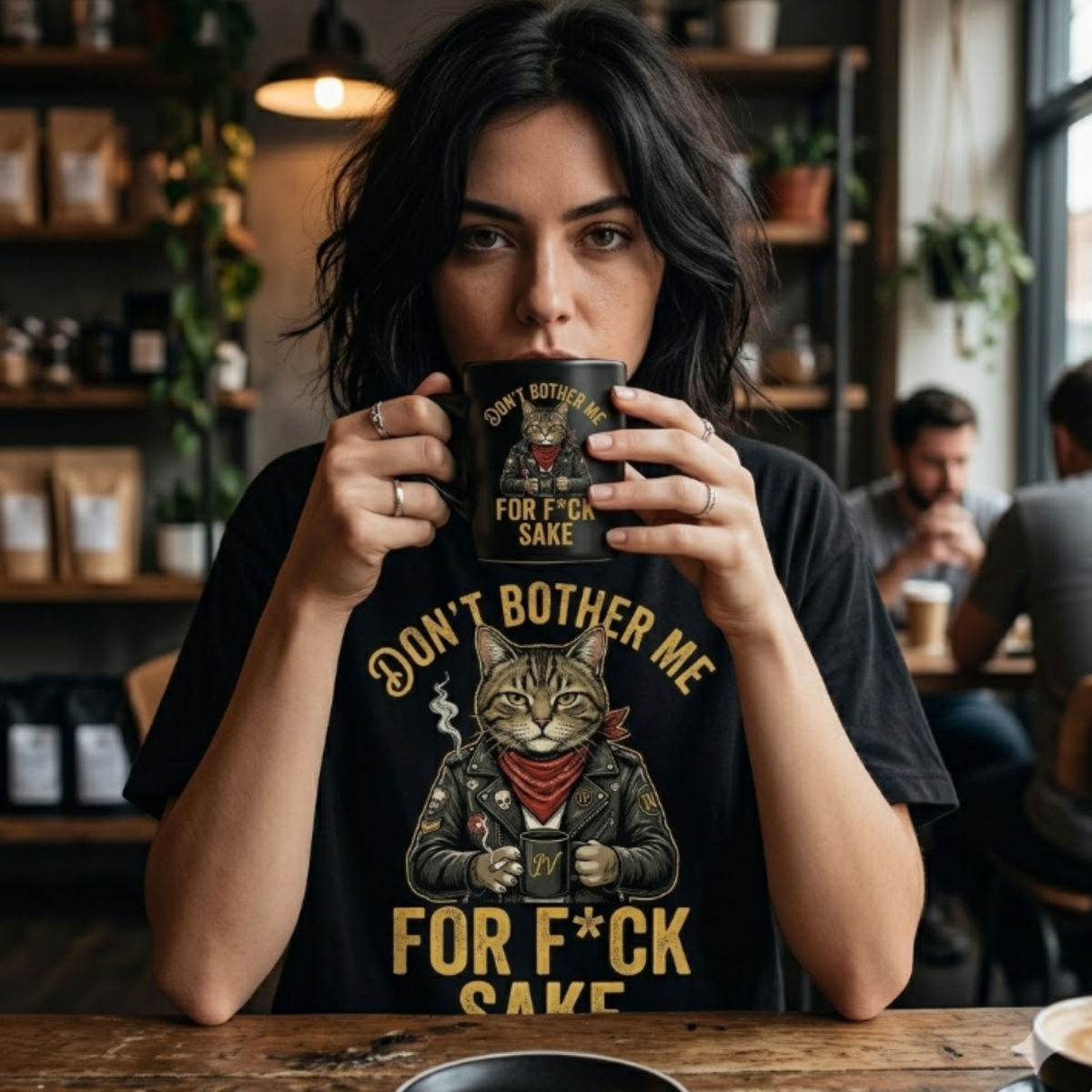 Person wearing a BIKER CAT tee that says 'Don't Bother Me' and 'FOR F*CK SAKE', holding a coffee mug in a café.